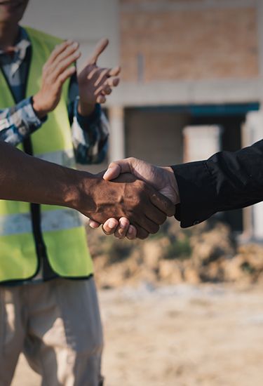 Duas pessoas apertam as mãos em um canteiro de obras. Uma, cliente Banco CNH, veste colete refletivo amarelo sobre camisa xadrez, enquanto a outra, de camisa preta, segura um laptop.