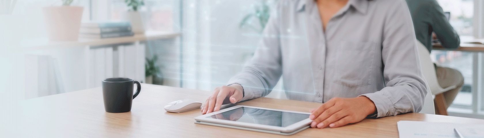 Mãos de uma mulher com camisa cinza ao lado de um tablet em uma mesa de madeira clara, com sutil interface holográfica. Uma caneca preta e um mouse branco completam o cenário de um escritório moderno.