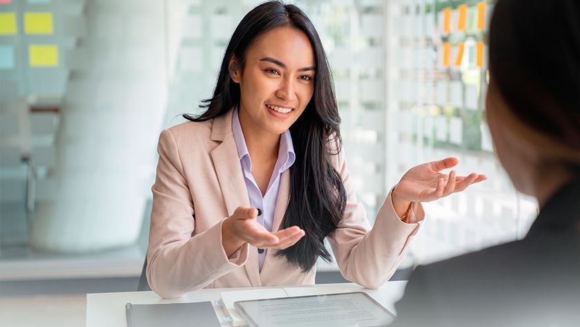 Uma mulher de longos cabelos escuros e blazer claro sorri e gesticula com as mãos enquanto conversa com outra pessoa sentada à sua frente. Ela está em uma mesa de escritório.