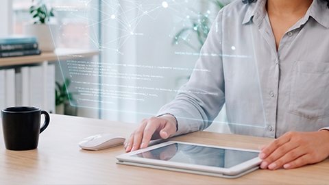 Mãos de uma mulher com camisa cinza ao lado de um tablet em uma mesa de madeira clara, com sutil interface holográfica. Uma caneca preta e um mouse branco completam o cenário de um escritório moderno.