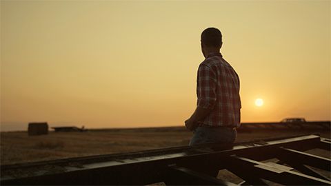 Homem vestindo camisa xadrez e calça jeans sentado sobre uma estrutura metálica agrícola. Ele observa um campo aberto sob a luz dourada de um pôr do sol.