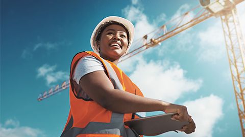 Mulher sorridente vestindo capacete branco e colete laranja segura um tablet. Ao fundo, há uma grua amarela de construção sob um céu azul ensolarado.