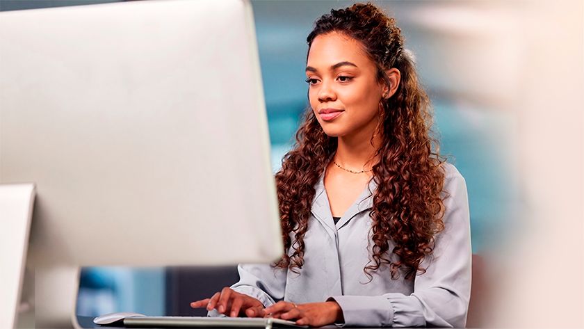 Uma mulher jovem de longos cabelos cacheados e camisa azul-clara digita concentrada em um computador de mesa. O fundo do escritório aparece desfocado em tons suaves de azul e branco.