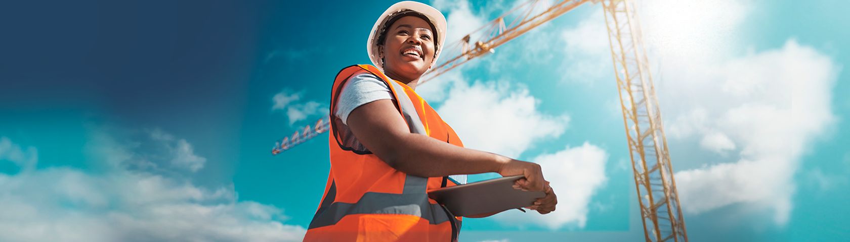 Uma mulher sorridente, vestindo capacete branco e colete laranja, segura um tablet em um canteiro de obras. Ao fundo, um guindaste amarelo se destaca sob um céu azul.