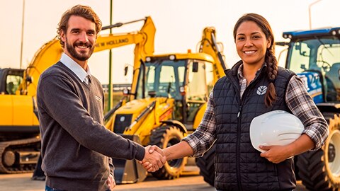 Um homem e uma mulher sorridentes apertam as mãos em frente a máquinas pesadas da New Holland amarelas e azuis.