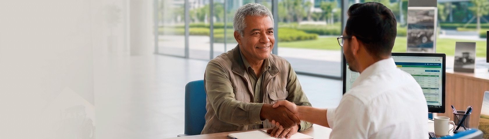 Um homem sorridente aperta a mão de outro homem em uma mesa de escritório. Ao fundo, através de um vidro com uma faixa azul, grandes máquinas agrícolas.