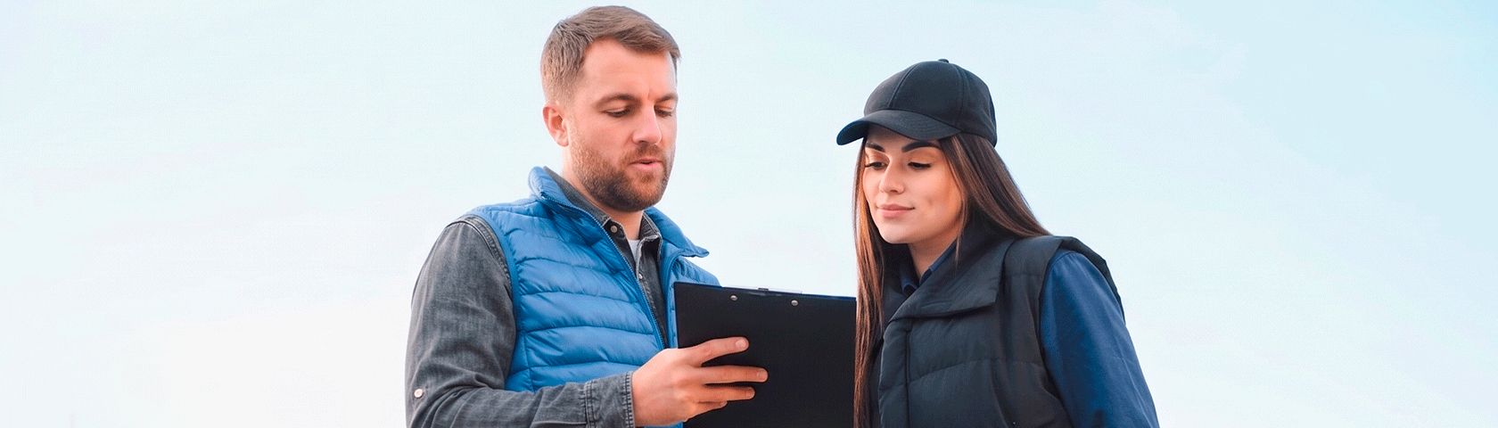 Um homem de colete azul e uma mulher de boné preto examinam juntos uma prancheta. Eles estão ao ar livre com um céu claro ao fundo.